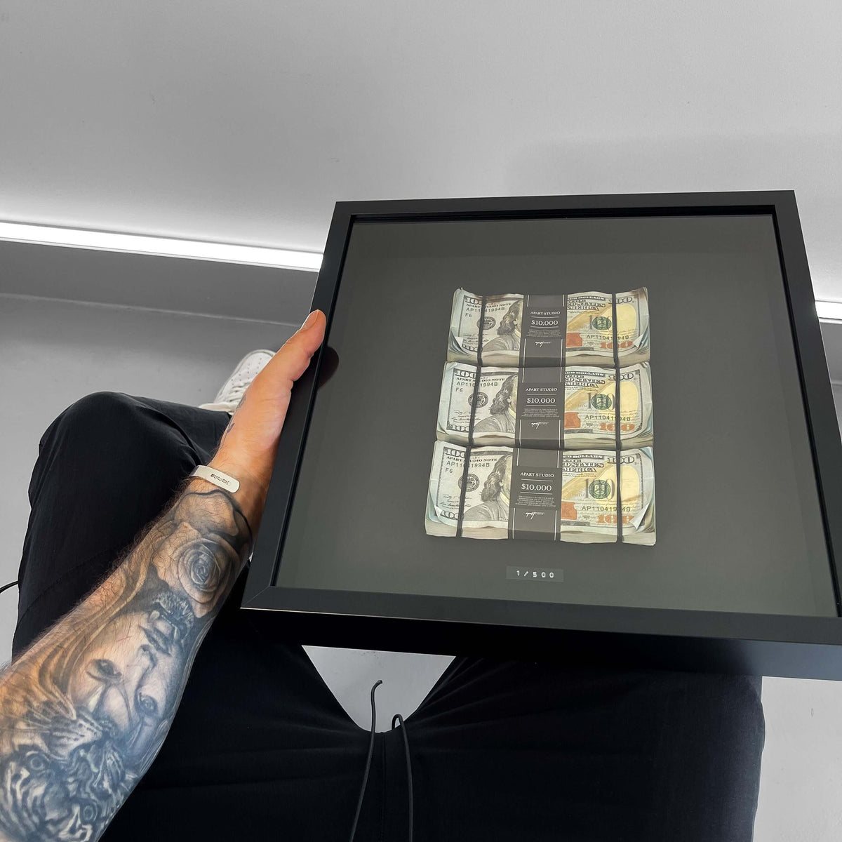 Framed artwork with printed money held by a person with tattoos.