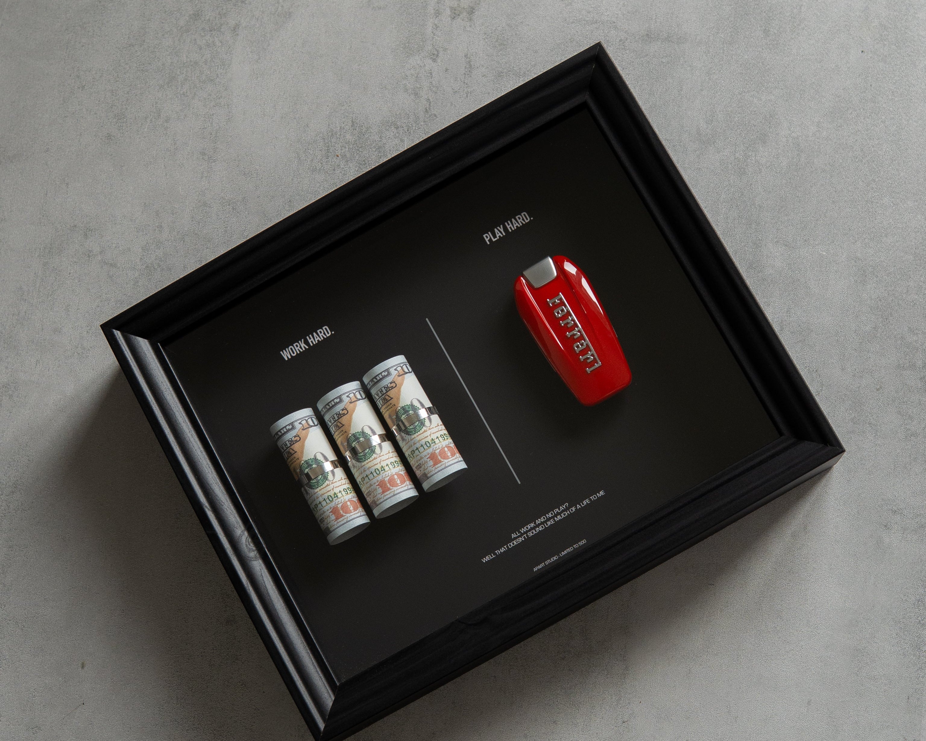 A black rectangular shadow box display on a gray concrete surface, containing a red Ferrari key fob, three rolled dollar bills, and printed text on a black background.