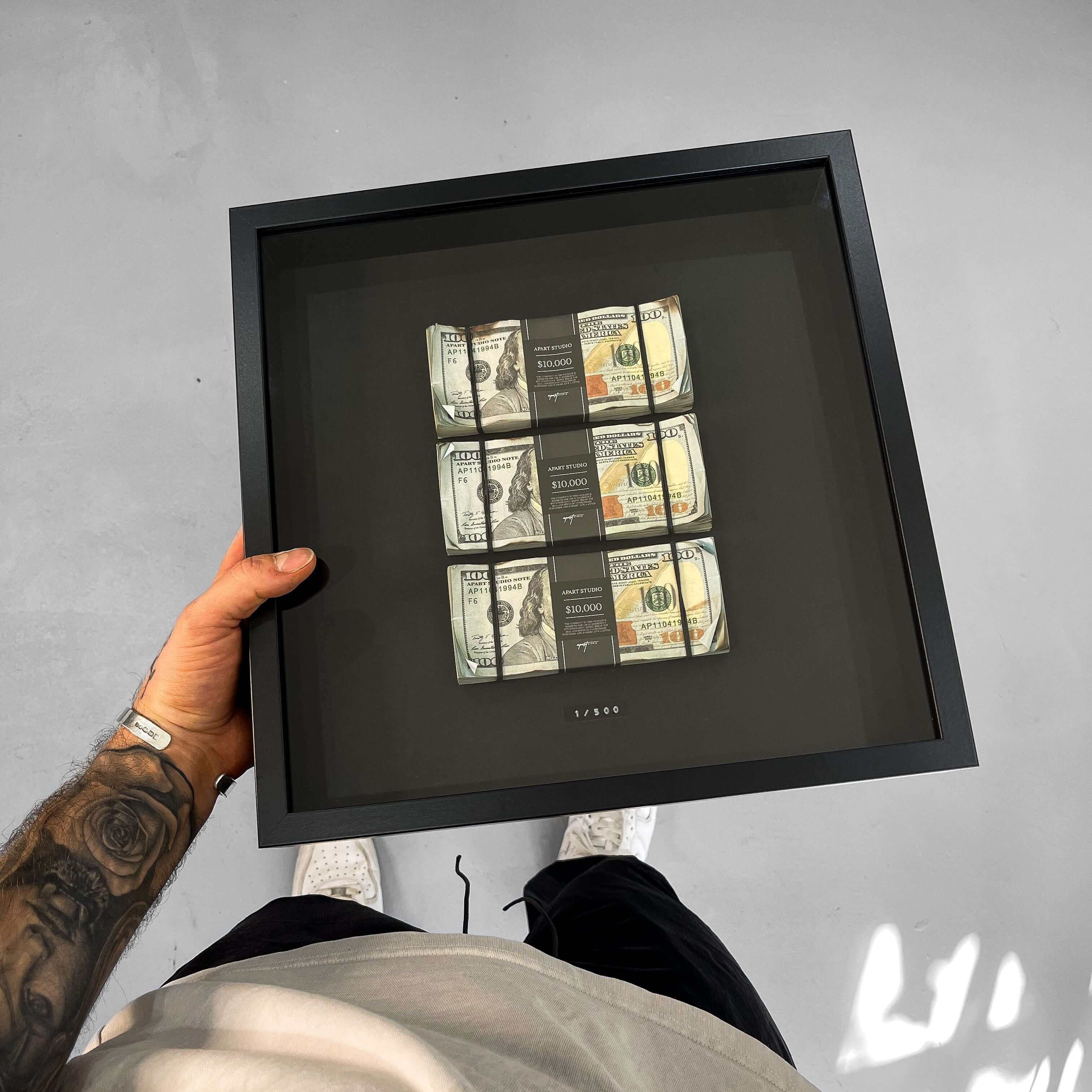 Framed artwork with dollar bills held by a person against a gray wall.