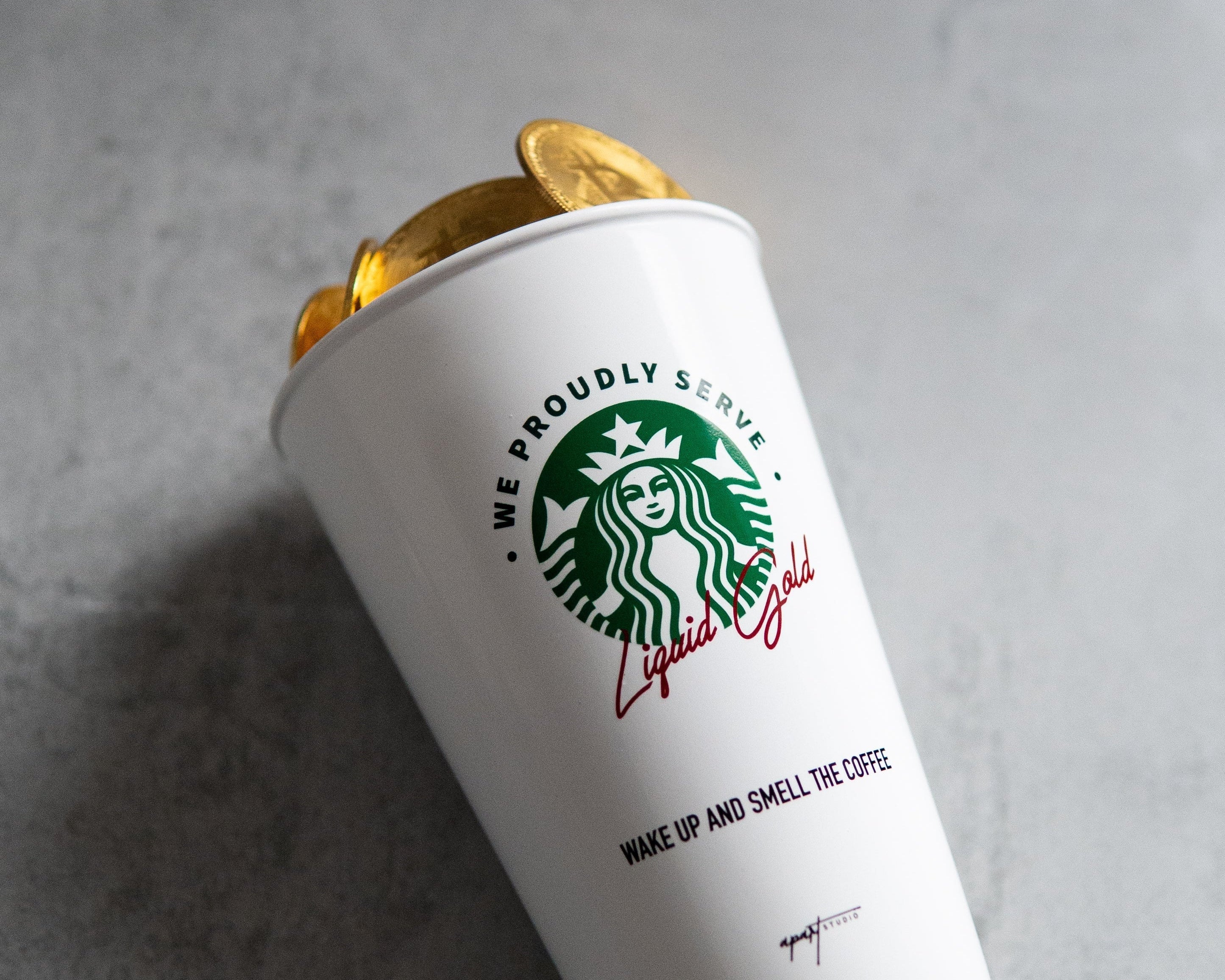 A white Starbucks paper coffee cup with a gold lid, photographed on a textured gray surface. The cup features the green Starbucks siren logo and the phrase 'We Proudly Serve' in green and red script.
